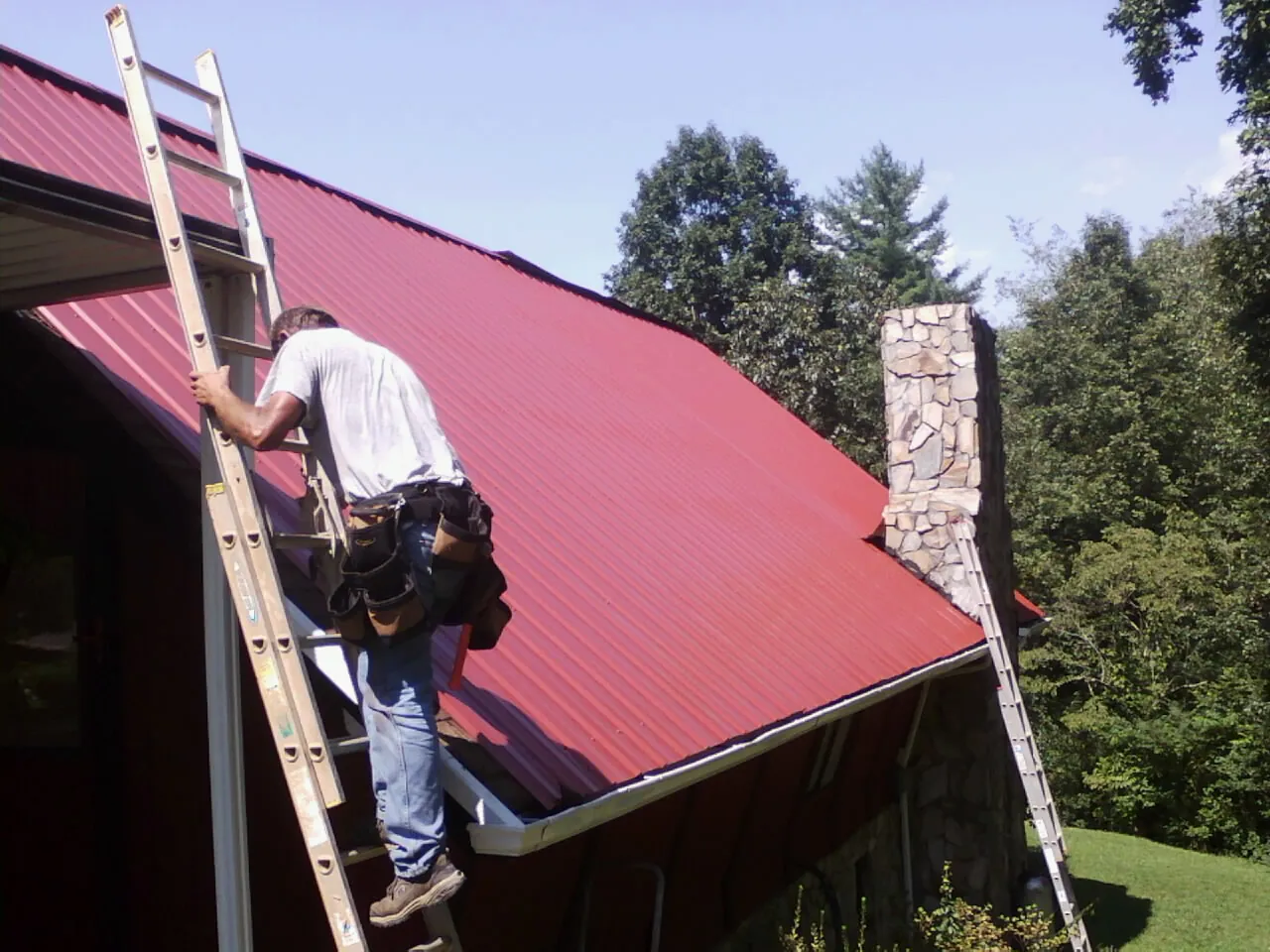 Roofer on a ladder beside a red metal roof and stone chimney — Soffit & Fascia Repair by Roofing Brothers in Hollis Center
