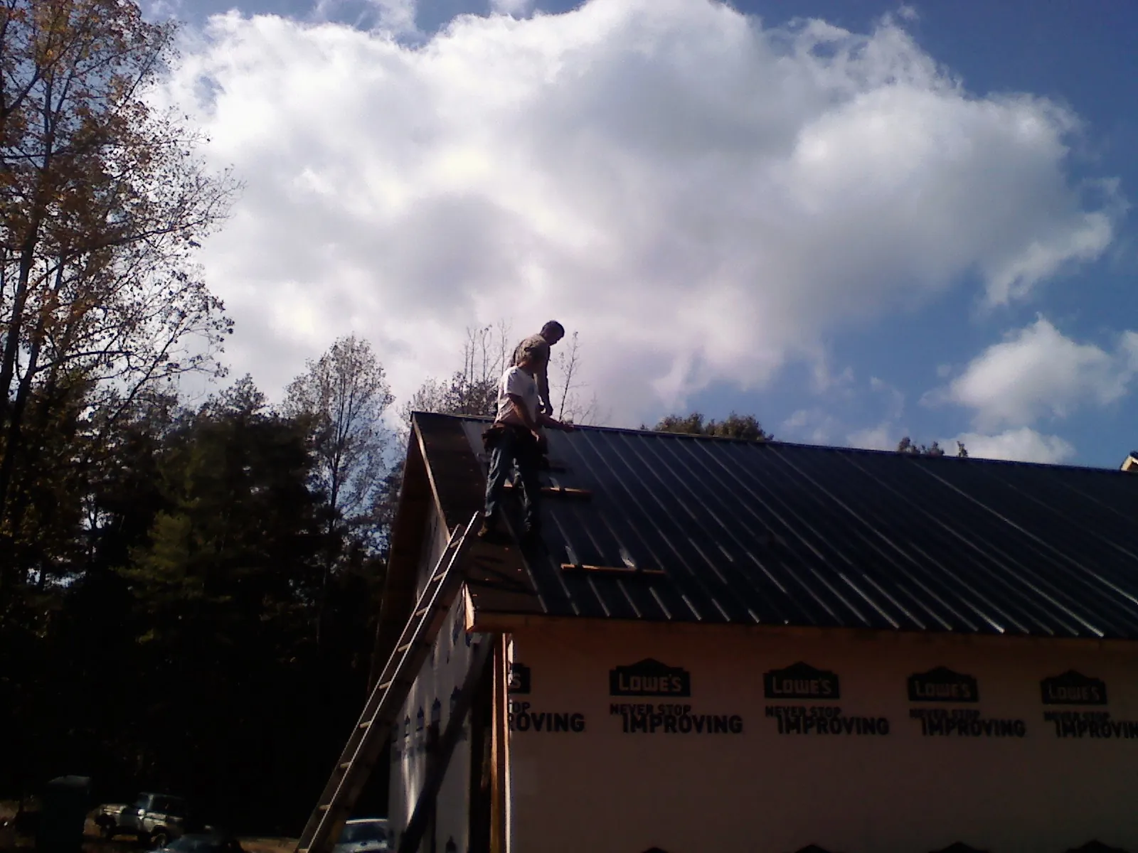 Roofer fastening dark metal panels high against a cloudy sky, from a Roof Inspection job in Hollis Center